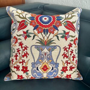 Puede incluir: Una almohada decorativa con fondo crema y un estampado floral colorido. El estampado presenta un jarr&oacute;n azul con flores rojas, amarillas y azules. La almohada est&aacute; sobre un sof&aacute; verde oscuro.