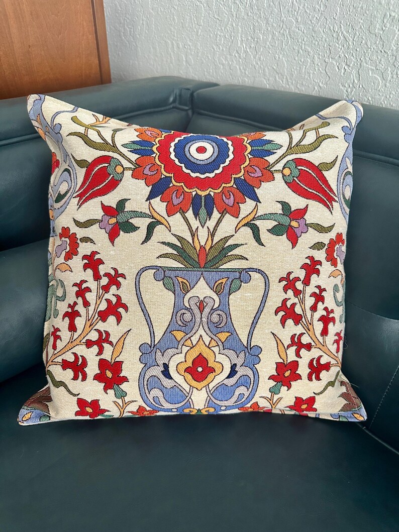 Puede incluir: Una almohada decorativa con fondo crema y un estampado floral colorido. El estampado presenta un jarr&oacute;n azul con flores rojas, amarillas y azules. La almohada est&aacute; sobre un sof&aacute; verde oscuro.