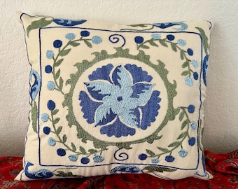 May include: A white throw pillow with blue and green embroidered floral designs. The pillow has a square shape and a cream-colored background.
