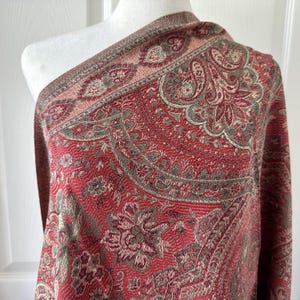 May include: A red paisley shawl with a detailed floral and scroll pattern. The shawl has a border of pink and green accents. The fabric is draped over a white mannequin, showcasing the intricate design and texture of the textile.