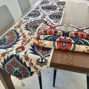 May include: A white table runner with a blue, red, and green floral pattern. The runner is folded in half and placed on a wooden table.