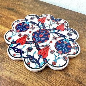 May include: A white, scalloped-edge ceramic coaster with a floral design. The design features red tulips, blue and red floral clusters, and blue stems and leaves. The coaster has a black border and is set on a wooden surface.
