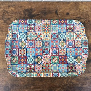May include: Rectangular serving tray with a colorful tile pattern. The tray features a repeating design of squares in shades of blue, red, yellow, and green. The edges of the tray are curved, and the tray is placed on a wooden surface.