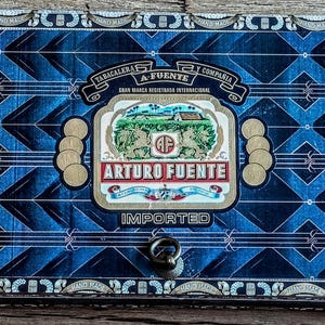 May include: A rectangular wooden box with a blue and black geometric pattern. The box features the text "ARTURO FUENTE IMPORTED" and a central emblem with a landscape scene and two lions. The box has a metal clasp.