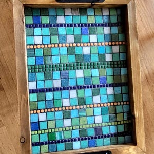 May include: A rectangular wooden serving tray with a mosaic tile design. The mosaic features a pattern of blue, green, and white square tiles, with orange and dark blue accents. The tray has black metal handles on each end.