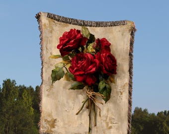 May include: A beige throw blanket featuring a painting of red roses. The blanket has a fringed edge and is held up against a blue sky and green trees. The roses are tied together with twine, creating a floral display.