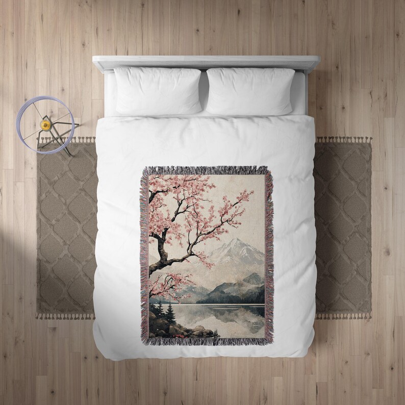 May include: A woven tapestry throw blanket featuring a watercolour-style painting of a cherry blossom tree with pink flowers, a mountain range, and a lake reflecting the sky. The throw blanket has a fringed border.