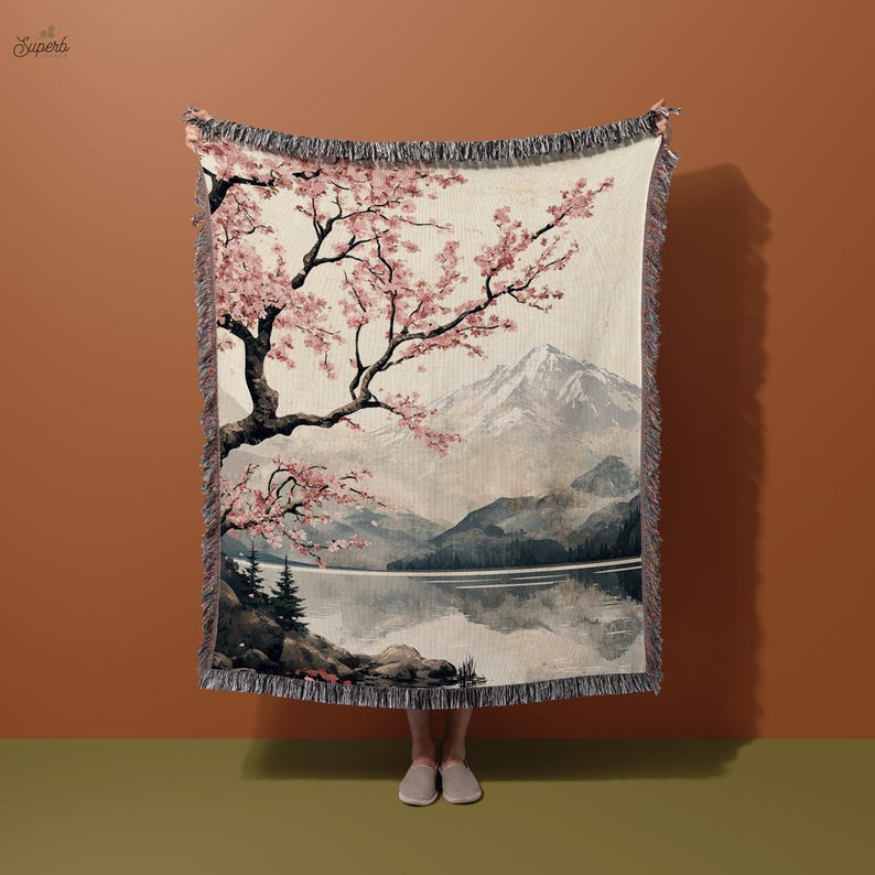 May include: A woven tapestry throw blanket featuring a scenic landscape design. The design depicts a mountain range, a lake, and a cherry blossom tree with pink flowers. The background is a soft beige colour.