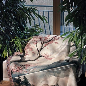May include: A woven tapestry throw blanket with a scenic landscape design featuring a cherry blossom tree, mountains, and a lake. The throw blanket has a fringe border and is draped over a chair.