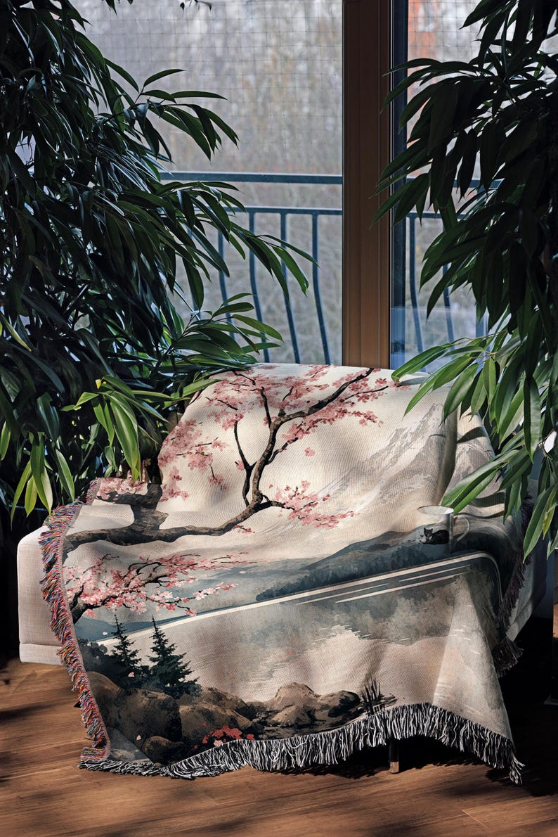 May include: A woven tapestry throw blanket with a scenic landscape design featuring a cherry blossom tree, mountains, and a lake. The throw blanket has a fringe border and is draped over a chair.