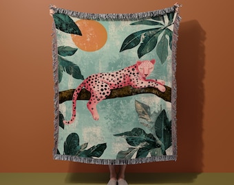 Op de afbeelding: Een roze en zwarte luipaardprint deken met een groen en bruin tropisch bladontwerp. De deken toont een luipaard dat slaapt op een tak met een grote oranje zon op de achtergrond.
