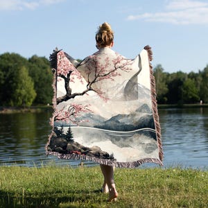 May include: A woman is holding a woven tapestry throw blanket with a scenic landscape design featuring a mountain range, a lake, and a cherry blossom tree. The throw blanket has a fringe border.