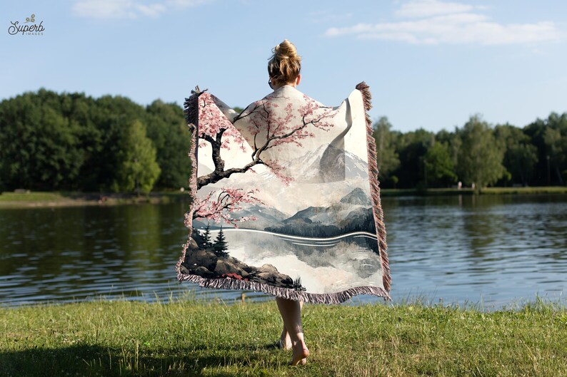 May include: A woman is holding a woven tapestry throw blanket with a scenic landscape design featuring a mountain range, a lake, and a cherry blossom tree. The throw blanket has a fringe border.