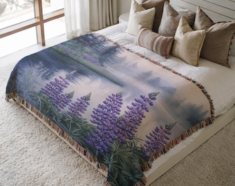 May include: A woven tapestry throw blanket features a scenic landscape with a lake and purple lupine flowers in the foreground. The blanket has a fringed edge and is displayed on a bed with pillows.