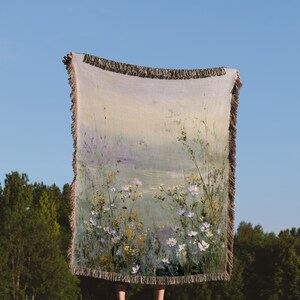 Puede incluir: Una manta rectangular con un diseño de paisaje floral, con flores blancas y moradas, se sostiene contra un cielo azul. La manta tiene un borde con flecos y una paleta de colores suaves, creando una estética serena y natural.