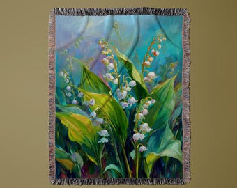May include: A woven throw blanket with a painting of lily of the valley. The artwork depicts white bell-shaped flowers, large green leaves, and a blue and green background. The blanket has a fringed edge.