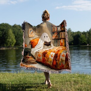 May include: A decorative throw blanket featuring a whimsical illustration of a ghost reading in bed. The blanket has a fringed edge and a warm color palette of orange, brown, and white, with a bookshelf and fall leaves design.