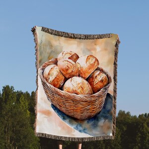 May include: A woven tapestry throw blanket with a watercolor painting of a wicker basket filled with freshly baked bread rolls.