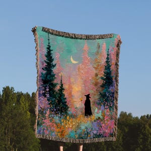 May include: A woven throw blanket featuring a colorful forest scene with a crescent moon and a silhouetted figure in a witch hat. The blanket has a fringed edge and is held up against a blue sky.