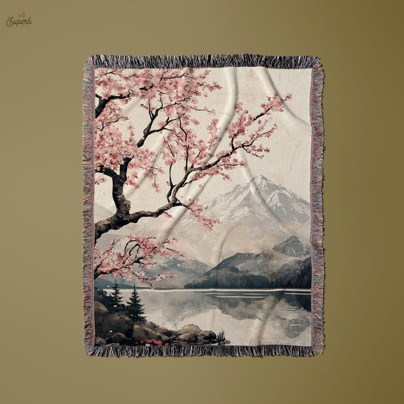 May include: A woven tapestry throw blanket featuring a watercolour-style painting of a cherry blossom tree with pink flowers, a mountain range, and a lake. The background is a light beige colour.