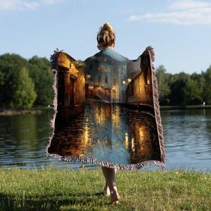 May include: A woven throw blanket featuring a nighttime cityscape scene with warm, golden light reflecting on water. The blanket has a fringed edge and is held up against a lake and green grass.