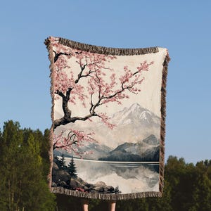 May include: A woven tapestry throw blanket featuring a Japanese-inspired landscape with a cherry blossom tree, a mountain, and a lake. The throw blanket has a fringe border.