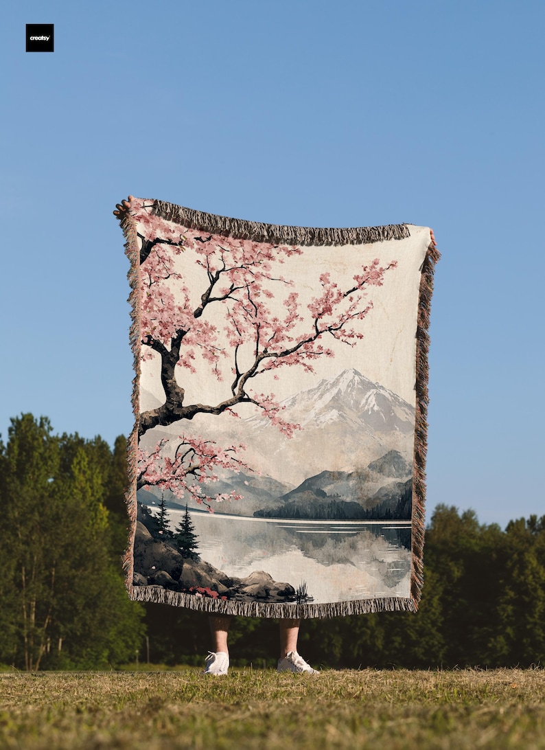 May include: A woven tapestry throw blanket featuring a Japanese-inspired landscape with a cherry blossom tree, a mountain, and a lake. The throw blanket has a fringe border.