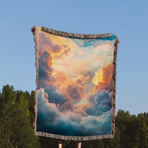 May include: A woven tapestry throw blanket with a colorful image of clouds and a sunset sky. The throw blanket has a fringe border.