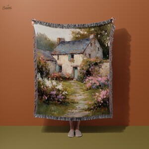 Puede incluir: Una manta tejida con una pintura de paisaje de una casa de piedra con tejado de pizarra, rodeada de vegetación y flores rosas. La obra de arte tiene un estilo vintage e impresionista con un borde con flecos.