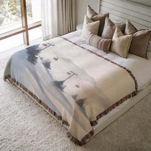 Può includere: Una coperta tessuta con una scena di seggiovia nei toni del blu, grigio e beige. La coperta ha un bordo con frange con un mix di colori. La coperta è drappeggiata su un letto con cuscini.