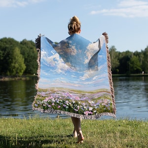 May include: A woven throw blanket with a landscape design featuring a blue sky, clouds, and a field of purple flowers. The blanket has a brown fringe and is held up by a person standing near a lake.