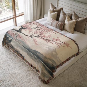 May include: A woven throw blanket with a cream background and a pink cherry blossom tree design. The tree is in front of a mountain range and a lake. The throw blanket is on a bed with pillows.