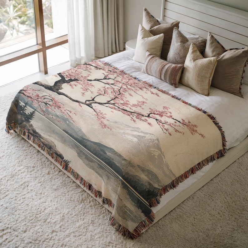 May include: A woven throw blanket with a cream background and a pink cherry blossom tree design. The tree is in front of a mountain range and a lake. The throw blanket is on a bed with pillows.