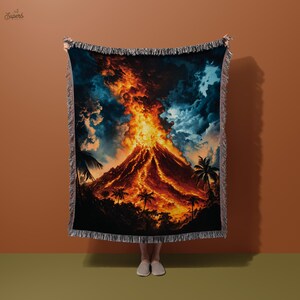May include: A woven throw blanket with a colorful image of a volcano erupting with lava and palm trees in the foreground. The blanket has a fringed border.