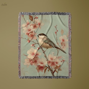 Puede incluir: Una manta rectangular con un pájaro posado en una rama floreciente. La manta presenta un fondo azul claro con una ilustración de estilo vintage de un pájaro y flores rosas. Los bordes están deshilachados.