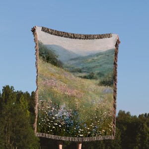 Puede incluir: Una manta tejida con una escena de paisaje de un campo de flores silvestres y colinas. La manta tiene un fondo crema con una imagen detallada de un prado en flor. Los bordes están acabados con una franja marrón.