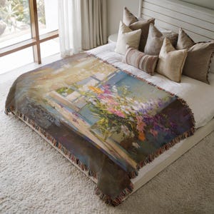 May include: A woven throw blanket featuring a floral still life painting in shades of blue, yellow, pink, and purple. The blanket has a fringed edge and is draped over a bed with pillows.