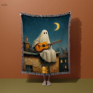 May include: A woven throw blanket featuring a whimsical illustration of a ghost playing a guitar on a rooftop under a starry night sky with a crescent moon. The artwork has a vintage, painterly style with warm tones.