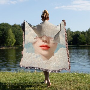 Puede incluir: Una manta tejida con un primer plano del rostro de una mujer, con labios rojo brillante y un fondo de cielo y nubes. La manta tiene un borde con flecos y la sostiene una persona cerca de un lago.