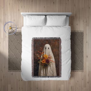 May include: A decorative throw blanket featuring a ghost holding a bouquet of flowers. The ghost is draped in a white sheet with two eye holes. The flowers are a mix of orange, yellow, and purple. The background is a dark brown.