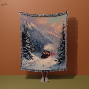May include: Woven blanket depicting a winter scene with a train in a snowy forest. The blanket has a fringed edge and shows a train with red carriages, snow-covered trees, and a mountain backdrop. The colour palette includes shades of blue, white, and red.