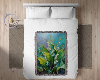 May include: A woven throw blanket with a floral painting of lily of the valley plants, with white bell-shaped flowers and large green leaves. The artwork has a blue and green background and a fringed border. The blanket is on a white bed.