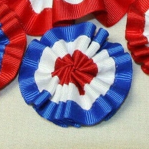 May include: Red, white, and blue fabric rosettes. The rosettes are made of ruffled ribbon and are arranged in a circular pattern.