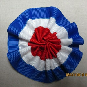 May include: A red, white, and blue rosette made of ribbon. The rosette is folded in a circular shape with the red ribbon in the center.