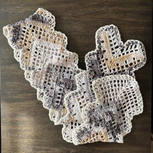 May include: A stack of handmade crochet coasters. Each coaster features a unique, openwork design with a scalloped edge. The color palette includes shades of cream, gray, and tan, creating a neutral and textured look.