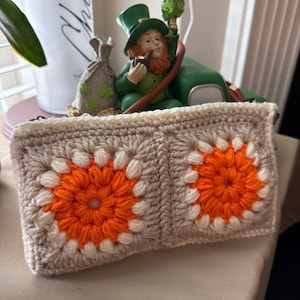 May include: A rectangular crochet pouch featuring two square motifs. Each motif has a bright orange center surrounded by a ring of cream yarn, set against a light beige background. The pouch is finished with a cream-colored border and a black zipper.