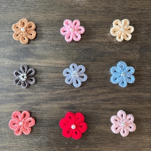 May include: Nine crocheted flower appliques in various colors, including tan, pink, and blue, are arranged on a wooden surface. Each flower features a central bead. The handmade floral embellishments are suitable for crafts and decorations.