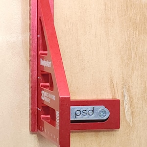 Peut inclure: Équerre de marquage 3D rouge de Woodpeckers, avec des mesures le long du bord. L'équerre porte le texte "psd" sur le côté et est posée sur un fond en bois clair. Un outil de précision pour le travail du bois.