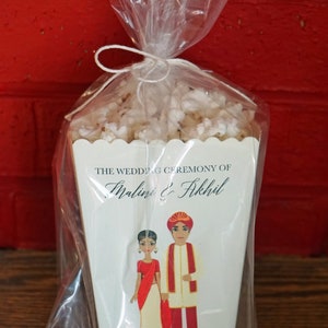 May include: A white favor box with a scalloped edge, filled with white popcorn. The box has a blue and black design with the text "The Wedding Ceremony of Malini & Akhil" and the date "21 January 2018, Rajkot, India".