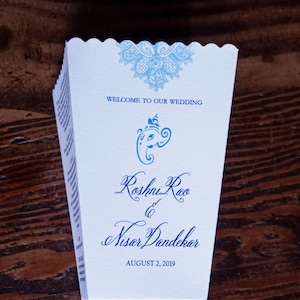 May include: A white wedding program with blue and white lace design. The program features the text "Welcome to our wedding" and the names "Roshni Rao & Nisar Dandekar" with the date "August 2, 2019".
